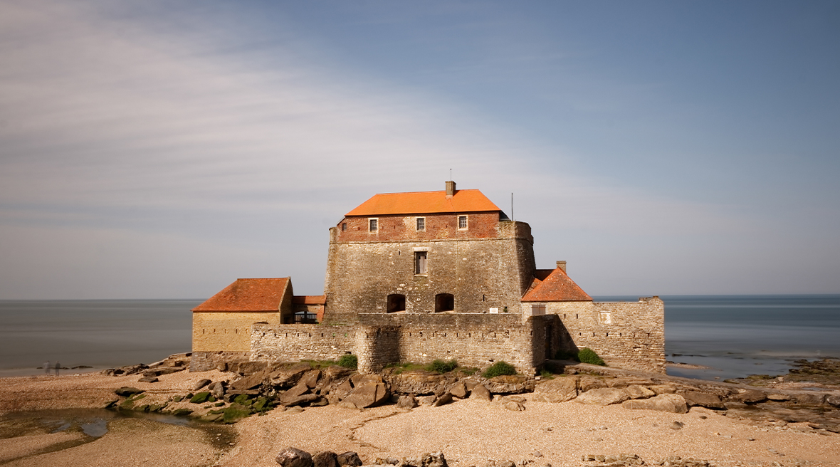 grey stone building with red tiled roof set on a sandy beach under blue skies